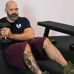 Bald man with beard doing Russian twists with a dumbbell, sitting on a gym floor.