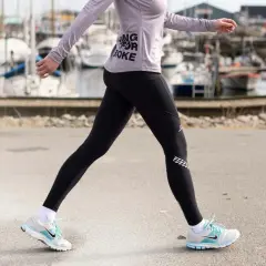 Kvinna i träningskläder går längs en hamn. Hon har på sig svarta tights och en lila tröja med texten "Running for Coke". Skorna är vita med blå detaljer. Hon tar många steg per km.