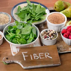 En skärbräda med texten "FIBER" omgiven av skålar med fiberrik mat: spenat, ärter, bönor, hallon, avokado, päron och spannmål.