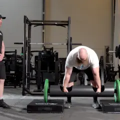 Man med skägg förbereder sig för farmers walk med en tung stock. En tränare ser på.