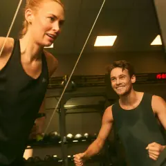 Två personer tränar på gym, en kvinna drar i ett rep och en man ler. De är fokuserade på att bygga muskler snabbt.