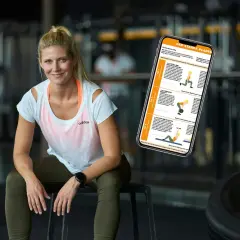 En kvinna sitter i ett gym med en telefon som visar en träningsplan. Hon ser ut att veta hur man blir stark.