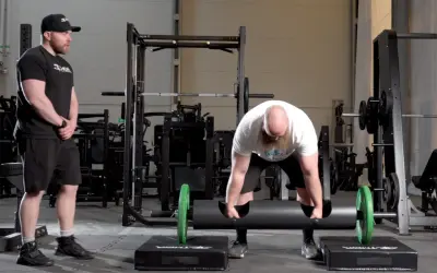 Man med skägg förbereder sig för farmers walk med en tung stock. En tränare ser på.