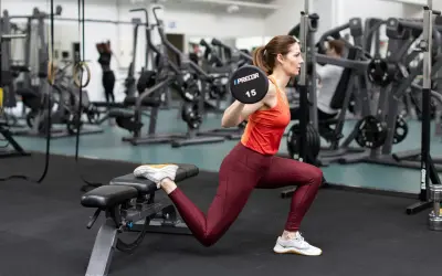 Kvinna utför bulgarian split squat med skivstång på axlarna i ett gym.