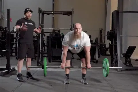 Instruktör visar rdl teknik för en man med skägg som förbereder sig för ett marklyft i ett gym.