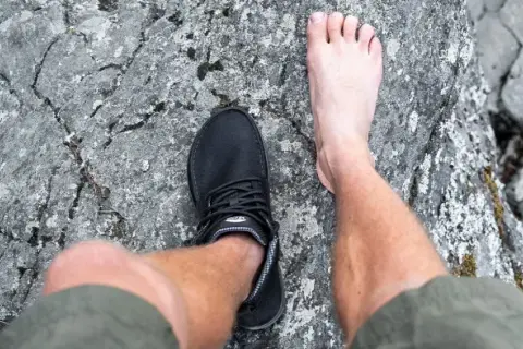 En barfota fot bredvid en barfotasko på en stenig yta. Frågan är om barfotaskor är bra eller dåligt för fötterna.