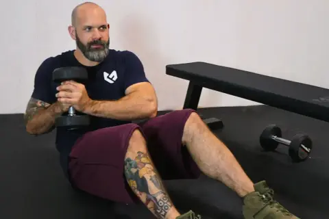 Bald man with beard doing Russian twists with a dumbbell, sitting on a gym floor.