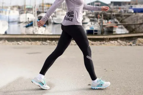 Kvinna i träningskläder går längs en hamn. Hon har på sig svarta tights och en lila tröja med texten "Running for Coke". Skorna är vita med blå detaljer. Hon tar många steg per km.