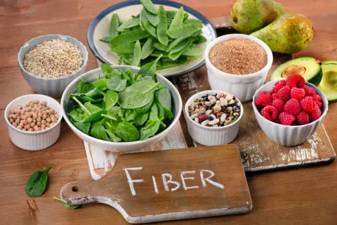 En skärbräda med texten "FIBER" omgiven av skålar med fiberrik mat: spenat, ärter, bönor, hallon, avokado, päron och spannmål.