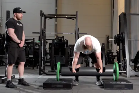 Man med skägg förbereder sig för farmers walk med en tung stock. En tränare ser på.
