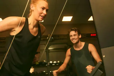Två personer tränar på gym, en kvinna drar i ett rep och en man ler. De är fokuserade på att bygga muskler snabbt.