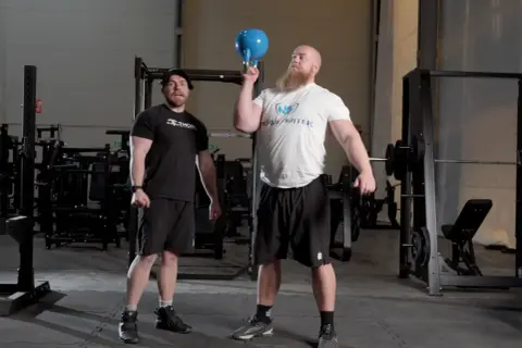 Man med skägg utför en kettlebell press med en blå kettlebell. En annan man står bredvid och tittar.