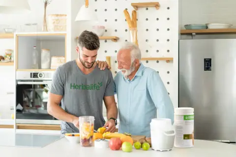 Två män i ett kök förbereder en smoothie med frukt. En burk med proteinpulver står på bänken, perfekt för att få rätt mängd protein per dag.