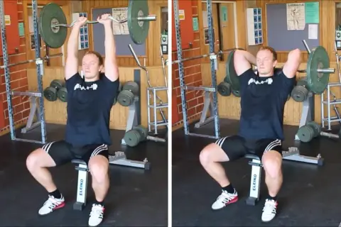 Man utför french press i gym. Han sitter på en bänk och lyfter en skivstång över huvudet.