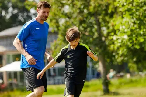 En man och en pojke springer på en gräsmatta, kanske på väg till ett **barn gym** eller en fotbollsträning.