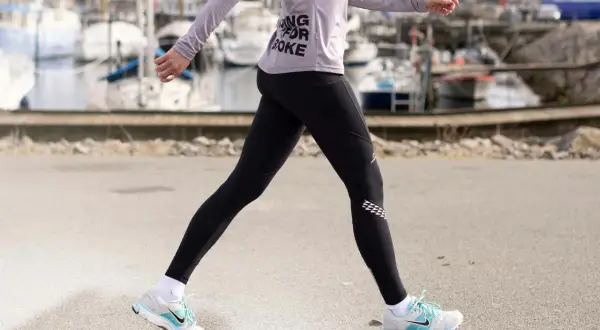 Kvinna i träningskläder går längs en hamn. Hon har på sig svarta tights och en lila tröja med texten "Running for Coke". Skorna är vita med blå detaljer. Hon tar många steg per km.