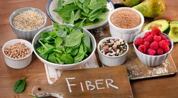 En skärbräda med texten "FIBER" omgiven av skålar med fiberrik mat: spenat, ärter, bönor, hallon, avokado, päron och spannmål.