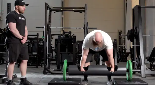 Man med skägg förbereder sig för farmers walk med en tung stock. En tränare ser på.