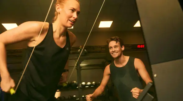Två personer tränar på gym, en kvinna drar i ett rep och en man ler. De är fokuserade på att bygga muskler snabbt.