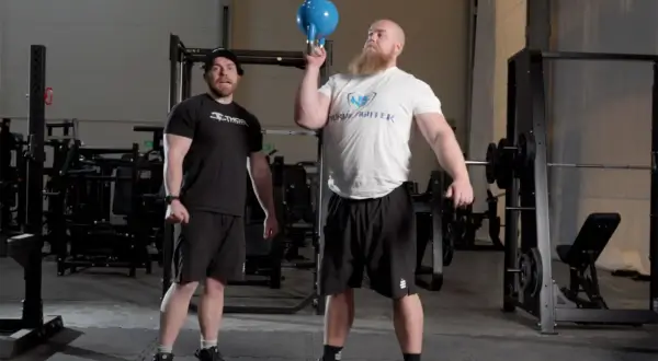 Man med skägg utför en kettlebell press med en blå kettlebell. En annan man står bredvid och tittar.