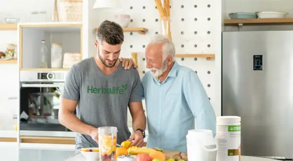 Två män i ett kök förbereder en smoothie med frukt. En burk med proteinpulver står på bänken, perfekt för att få rätt mängd protein per dag.