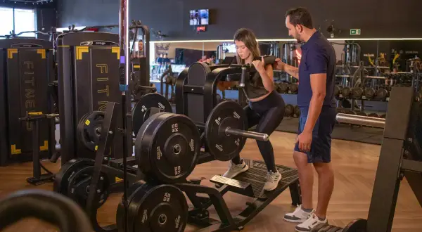 Kvinna utför benpress med instruktör bredvid. En del av ett gym med vikter och maskiner, perfekt för en bro split.