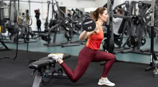 Kvinna utför bulgarian split squat med skivstång på axlarna i ett gym.