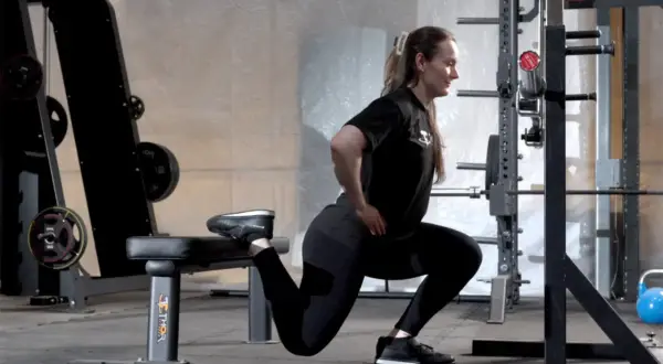 Kvinna utför split squats i ett gym. Hennes ena fot vilar på en bänk bakom henne medan hon böjer på det främre benet.