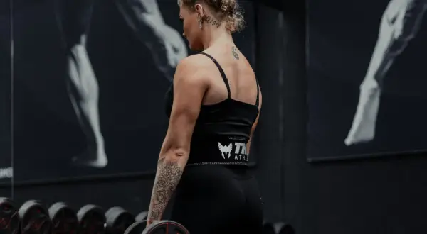 Kvinna med tatueringar utför shrugs med hantlar i ett gym.