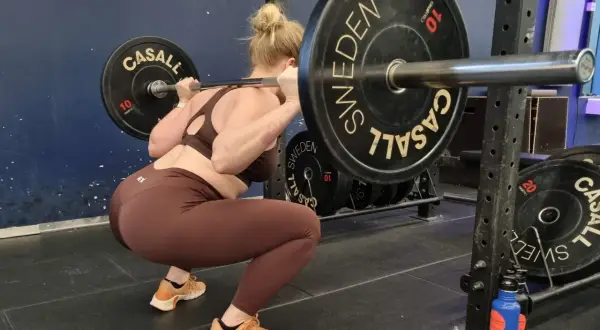 Kvinna gör squats med viktstång på axlarna. Träningspasset är i full gång med tunga viktskivor märkta "SWEDEN".