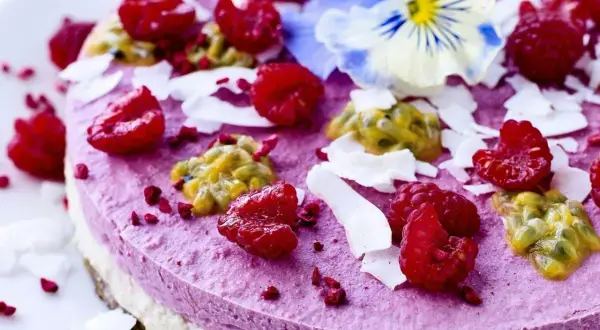 En vacker, lila tårta toppad med hallon, passionsfrukt, kokosflingor och en blå blomma. Perfekt för dig som söker nyttiga kakor.