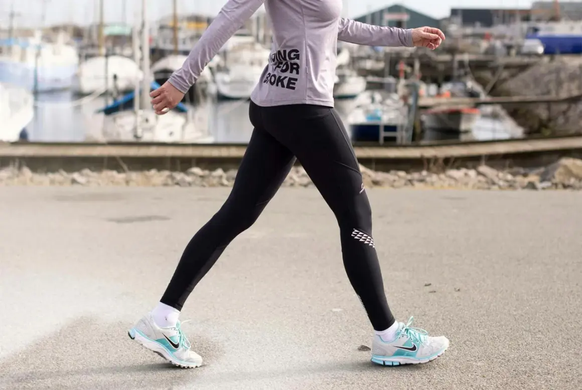 Kvinna i träningskläder går längs en hamn. Hon har på sig svarta tights och en lila tröja med texten "Running for Coke". Skorna är vita med blå detaljer. Hon tar många steg per km.