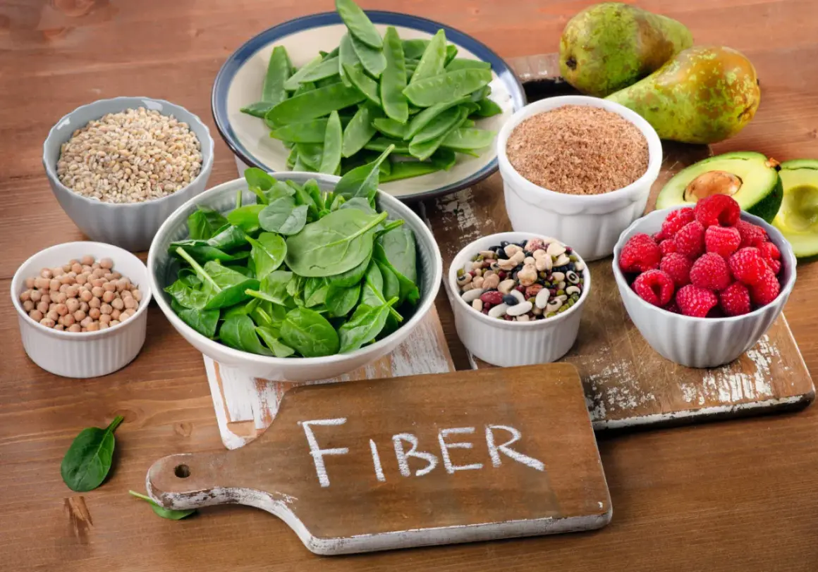 En skärbräda med texten "FIBER" omgiven av skålar med fiberrik mat: spenat, ärter, bönor, hallon, avokado, päron och spannmål.