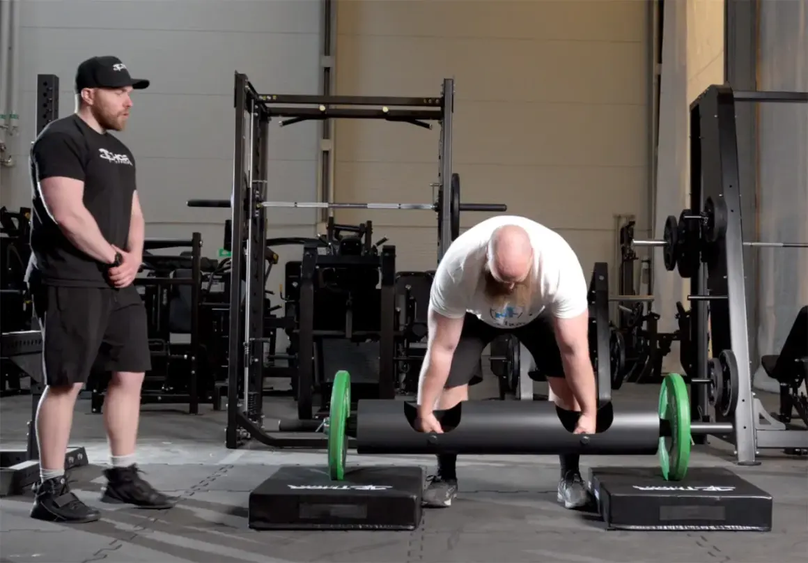Man med skägg förbereder sig för farmers walk med en tung stock. En tränare ser på.