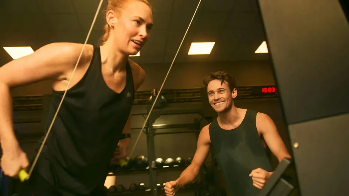 Två personer tränar på gym, en kvinna drar i ett rep och en man ler. De är fokuserade på att bygga muskler snabbt.