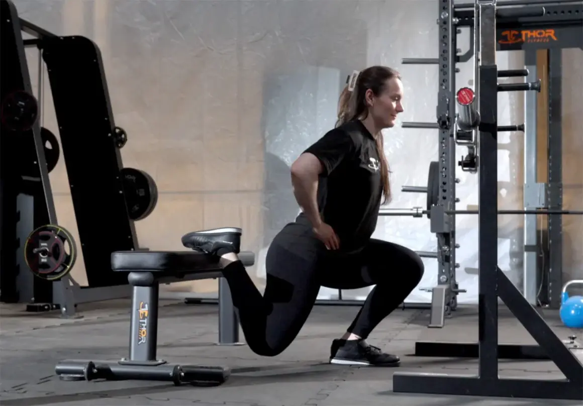 Kvinna utför split squats i ett gym. Hennes ena fot vilar på en bänk bakom henne medan hon böjer på det främre benet.