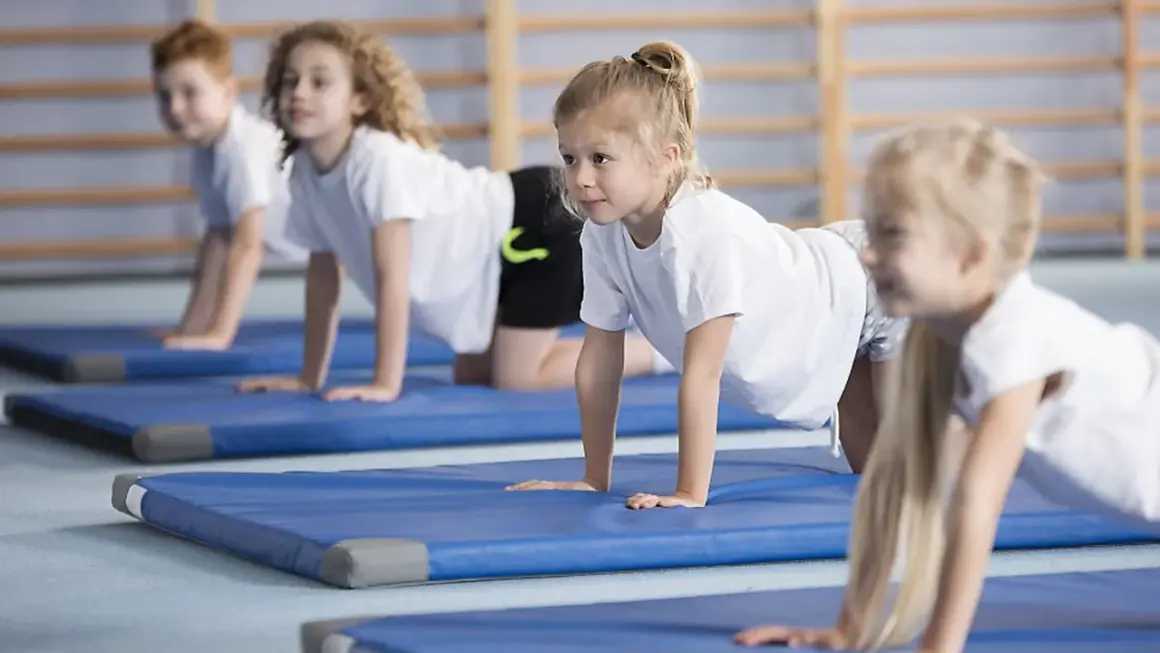 Barn på gympaövning, en aktivitet som motverkar stillasittande barn. De ligger på blåa mattor i en gymnastikhall.