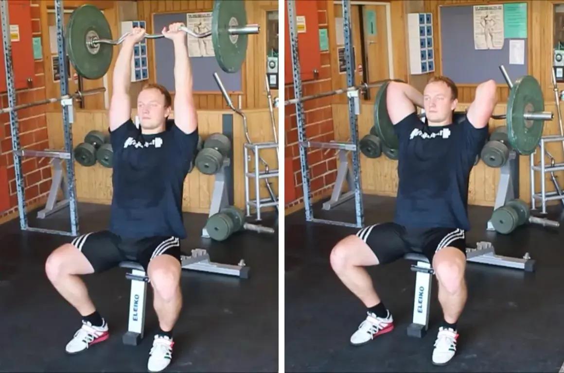 Man utför french press i gym. Han sitter på en bänk och lyfter en skivstång över huvudet.