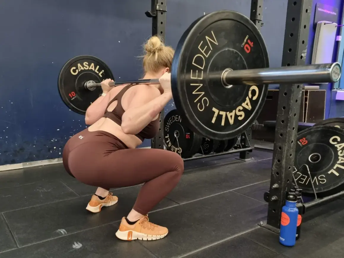 Kvinna gör squats med viktstång på axlarna. Träningspasset är i full gång med tunga viktskivor märkta "SWEDEN".