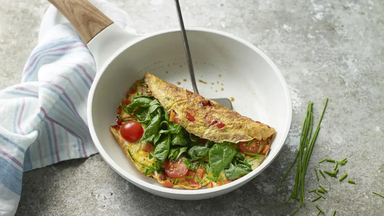 En gyllene, nyttig omelett fylld med spenat, k&ouml;rsb&auml;rstomater och paprika, serverad i en vit panna med tr&auml;handtag.
