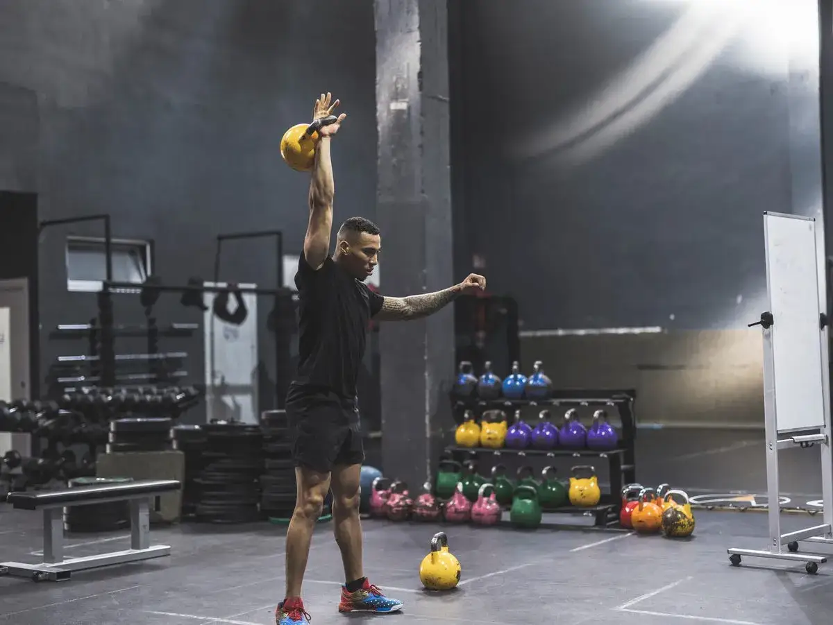 Man utf&ouml;r en kettlebell pass med en gul kettlebell &ouml;ver huvudet i ett gym.