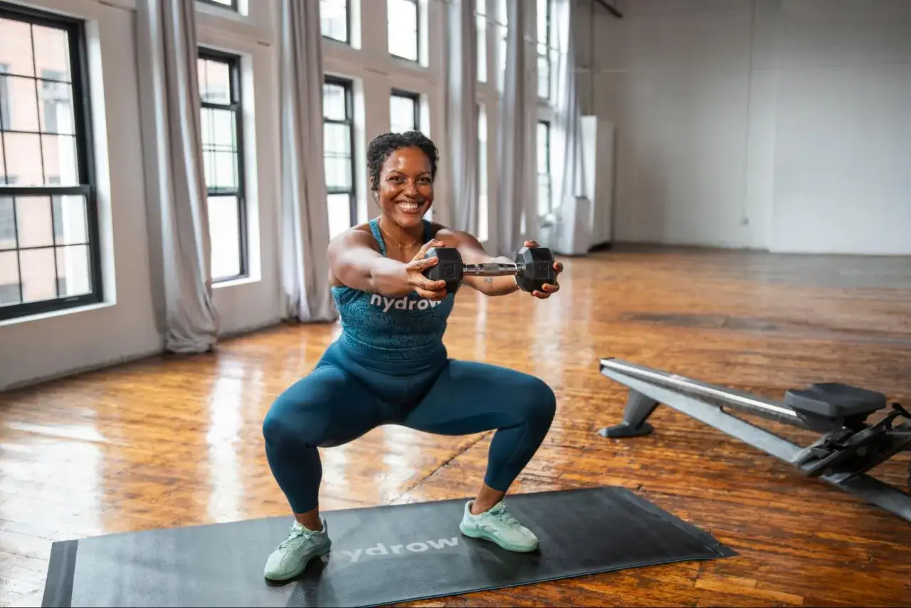 Kvinna utf&ouml;r helkroppspass gym med hantlar i en ljus lokal med stora f&ouml;nster.