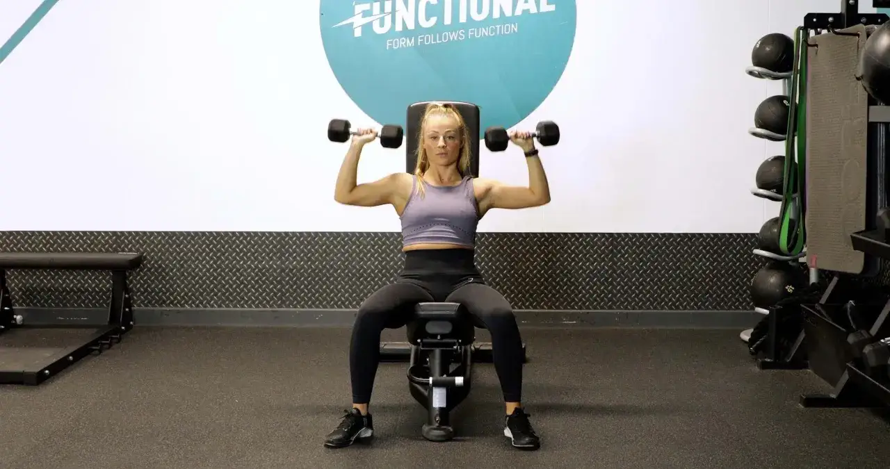 Kvinna utf&ouml;r sittande axelpress med hantlar i ett gym.