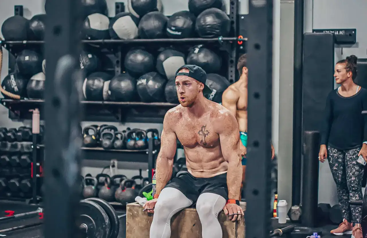 En svettig man med en imponerande crossfit kropp vilar p&aring; en box. I bakgrunden syns kettlebells och medicinbollar.
