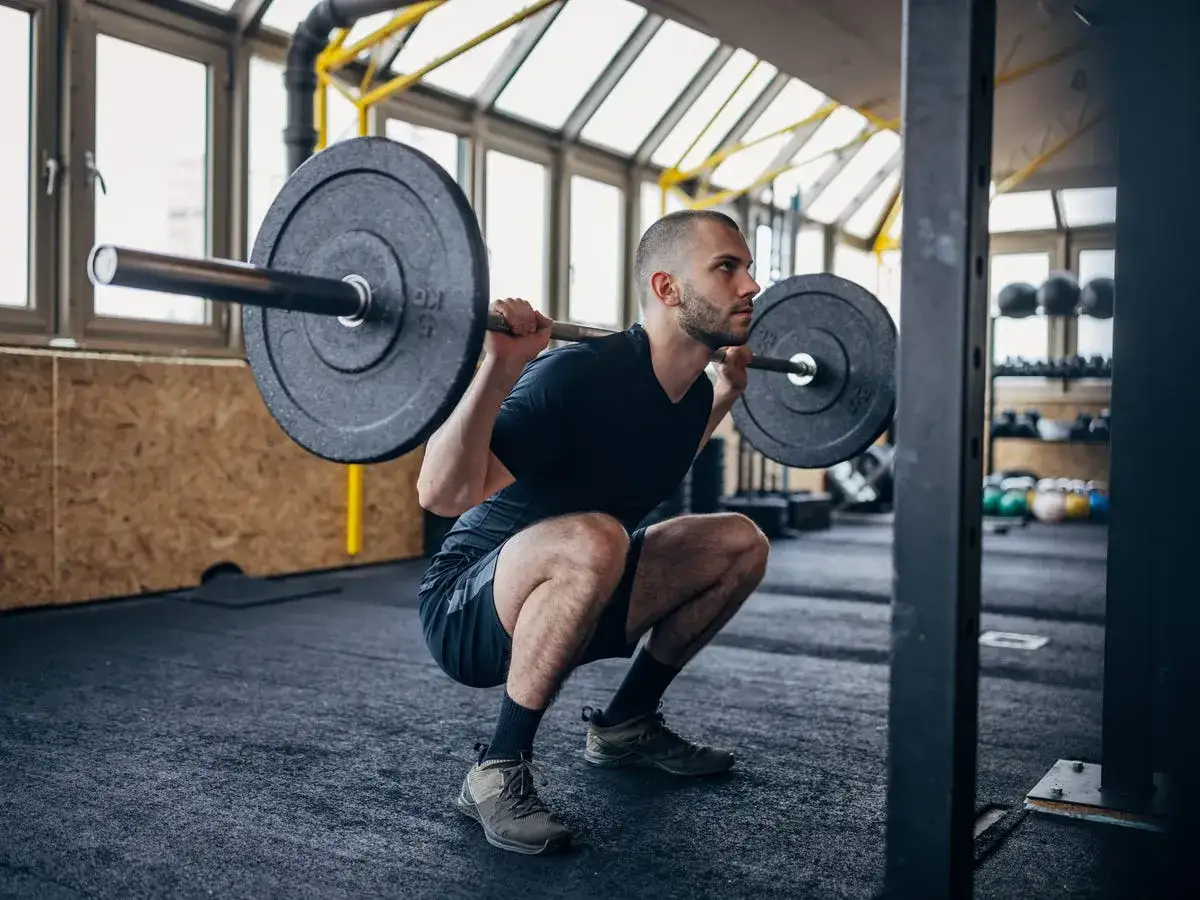 Man g&ouml;r squats med vikt i ett gym. Han &auml;r fokuserad och har en skivst&aring;ng p&aring; axlarna.