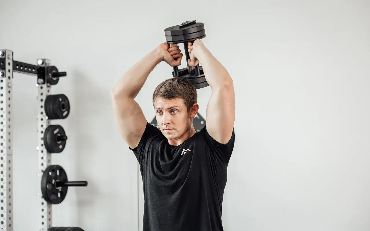 Man utför en french press i gymmet med en hantel. Viktskivor syns i bakgrunden.