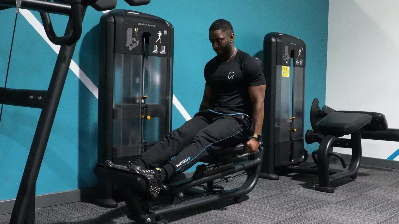 Man tränar sittande vadpress på ett gym. Maskinen är svart och har en blå vägg bakom sig.