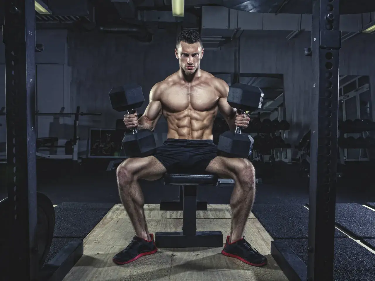 Muskul&ouml;s man med ektomorf kroppstyp sitter p&aring; en b&auml;nk i ett gym och lyfter hantlar.