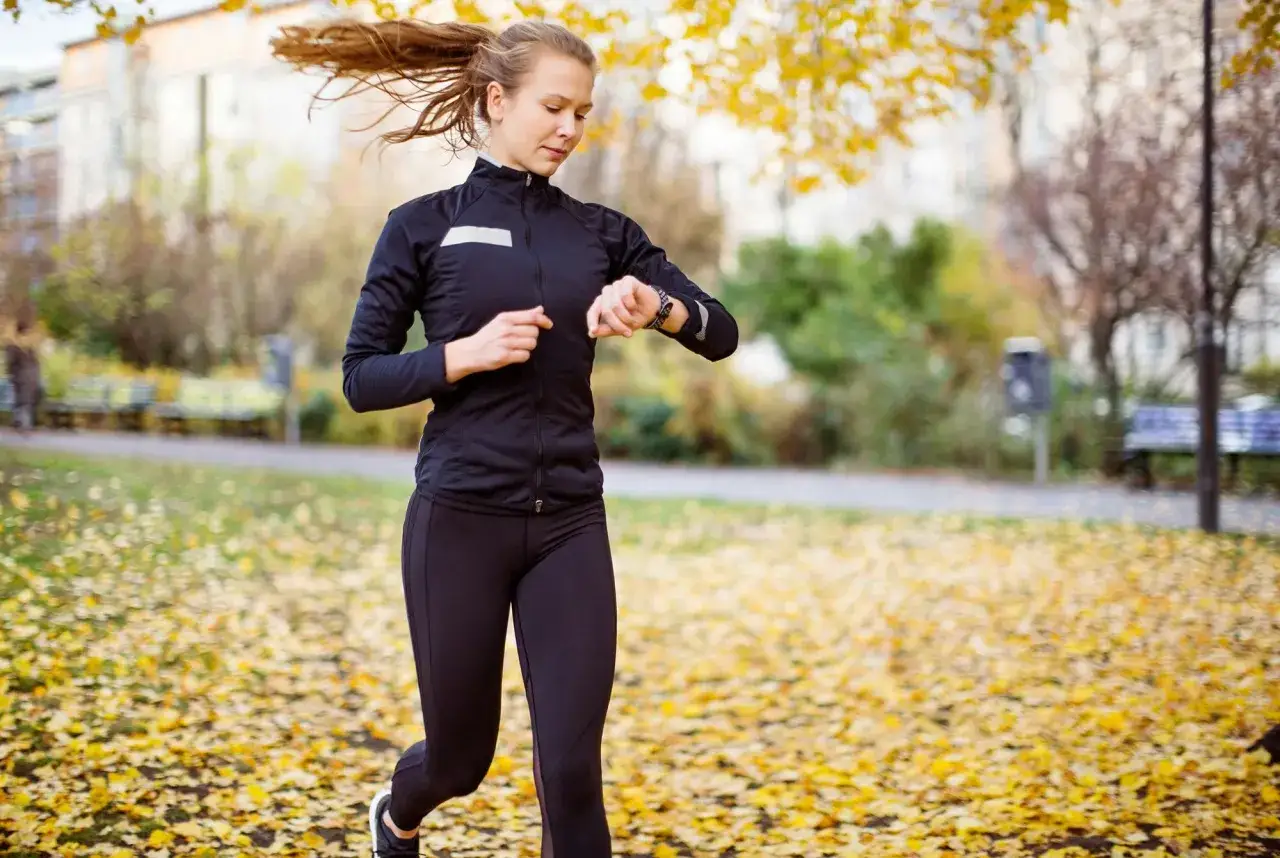 En kvinna springer i en park t&auml;ckt av h&ouml;stl&ouml;v och kollar sin klocka, hon verkar ha en bra tid p&aring; 5 km.