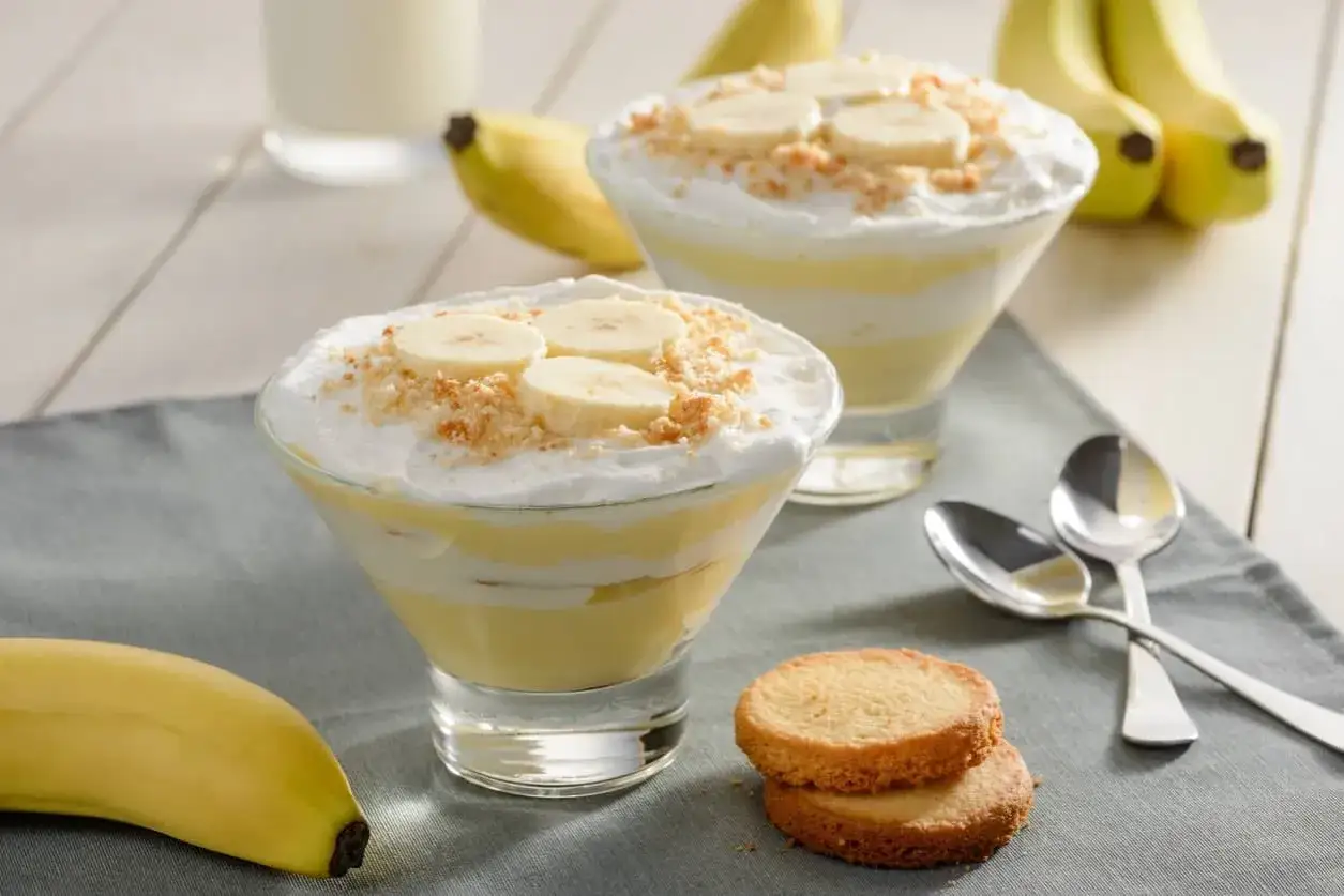 Två glas med bananpudding, toppad med grädde, kexsmulor och bananskivor. En banan ligger bredvid. Banan nyttigt!